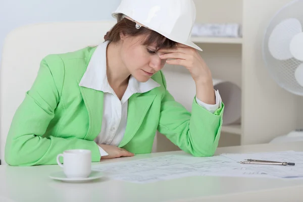 Tired woman - engineer in helmet sits at table i