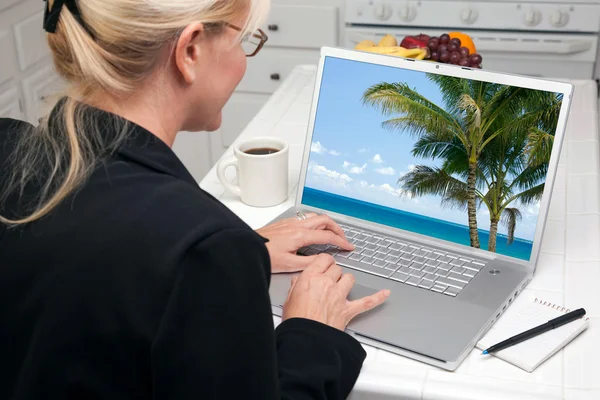 Woman In Kitchen Using Laptop to Research Travel with Palm Trees on Screen