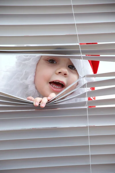 Little girl Santa Claus looking to snow through the blinds — Stock Photo, Image
