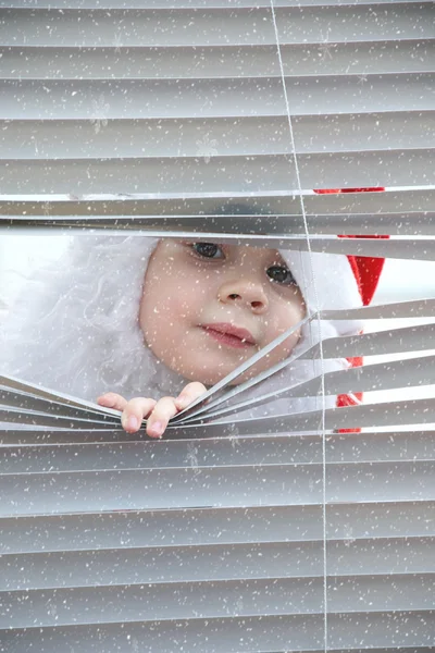 Klein meisje santa claus op zoek naar sneeuw via de blinds aparting hen met handen — Stockfoto