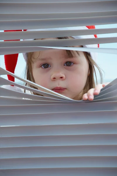 Little girl Santa Claus looking to snow through the blinds aparting them with hands — Stock Photo, Image