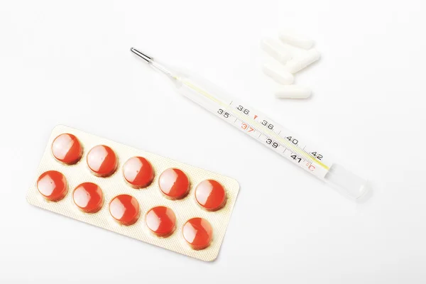 Termômetro entre um pacote de comprimidos vermelhos e cápsulas brancas — Fotografia de Stock