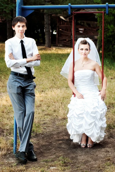 Bride sitting on swings and groom standing near — Stock Photo, Image