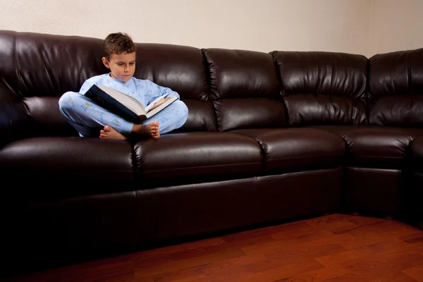 Miúdo bonito lendo um grande livro em um sofá — Fotografia de Stock