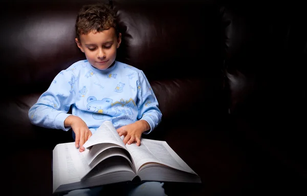 Miúdo bonito lendo um grande livro em um sofá — Fotografia de Stock