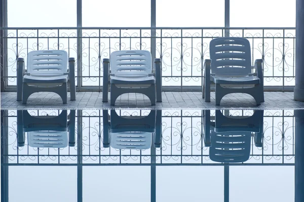 The plastic reclining chairs before the glass window pane by the swimming pool.
