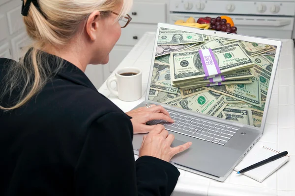 Жінка на кухні використовуючи ноутбук зі стосами грошей на екрані — стокове фото