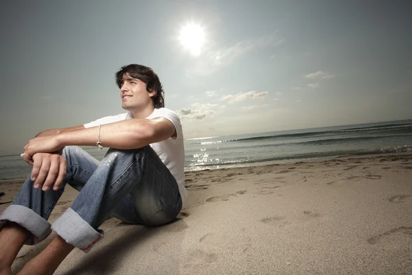 男子坐在与太阳在他身后的沙滩上 — 图库照片